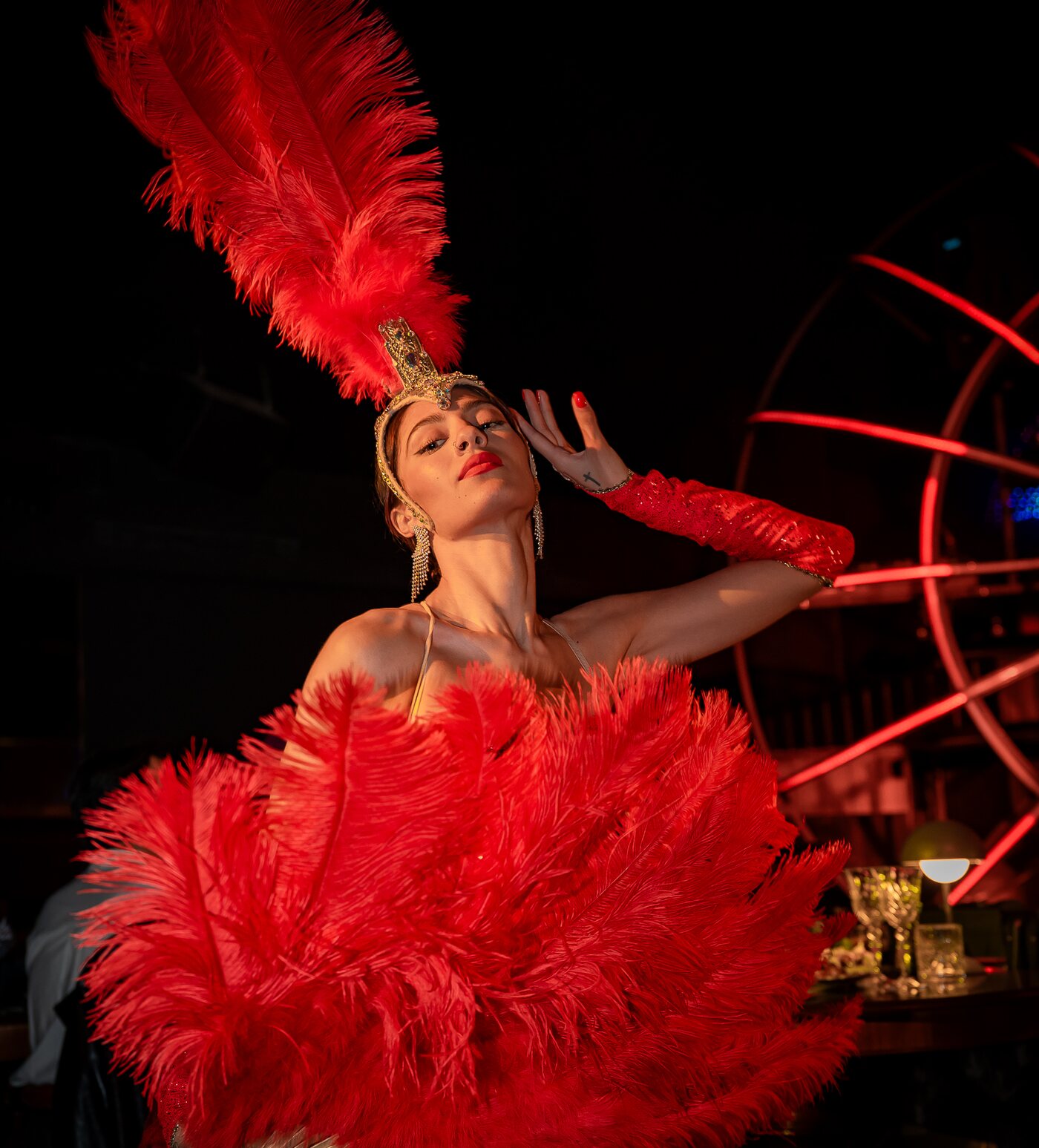 Performer con copricapo e abito di piume rosse accanto ai tavoli, davanti al cerchio di neon